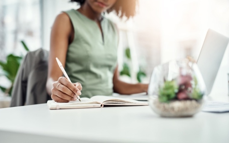 A person writing in a notebook beside an open laptop